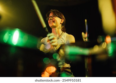 Female drummer playing drums with drumsticks and smiling on stage during a live performance, illuminated by colorful stage lights - Powered by Shutterstock