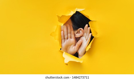 Female ear and hands close-up. Copy space. Torn paper, yellow background. The concept of eavesdropping, espionage, gossip and the yellow press. - Shutterstock ID 1484273711