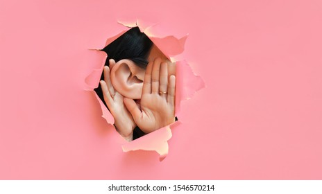 Female ear and hands close-up. Copy space. Torn paper, pink background. The concept of eavesdropping, espionage, gossip and the yellow press. Caricature with an enlarged ear.