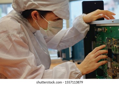 A Female Electronic Manufacturing Employee With Clean Suit (smock), Cap And Mask Inspecting A Circuit Board.