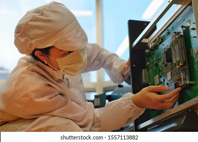 A Female Electronic Manufacturing Employee With Clean Suit (smock), Cap And Mask Inspecting A Circuit Board.