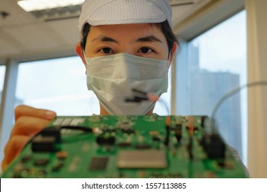 A Female Electronic Manufacturing Employee With Clean Suit (smock), Cap And Mask Inspecting A Circuit Board.