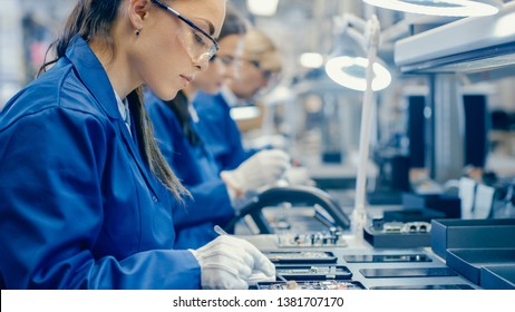 Female Electronics Factory Workers in Blue Work Coat and Protective Glasses Assembling Printed Circuit Boards for Smartphones with Tweezers. High Tech Factory with more Employees in the Background. - Shutterstock ID 1381707170