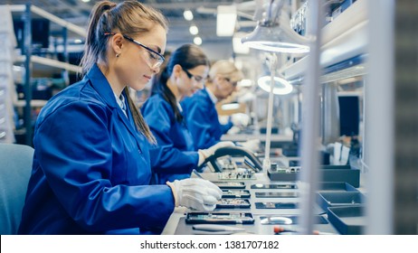 Female Electronics Factory Workers in Blue Work Coat and Protective Glasses Assembling Printed Circuit Boards for Smartphones with Tweezers. High Tech Factory with more Employees in the Background. - Shutterstock ID 1381707182