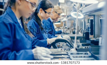 Female Electronics Factory Workers in Blue Work Coat and Protective Glasses Assembling Printed Circuit Boards for Smartphones with Tweezers. High Tech Factory with Employees.