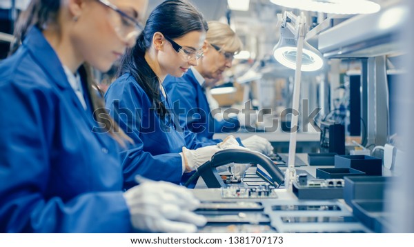 Female
Electronics Factory Workers in Blue Work Coat and Protective
Glasses Assembling Printed Circuit Boards for Smartphones with
Tweezers. High Tech Factory with
Employees.