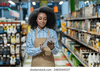 Female employee in a retail store aisle, using a tablet for inventory management. She wears a checkered shirt and an apron, focusing on digital tasks surrounded by various products. - Powered by Shutterstock