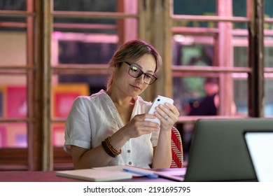 Female Employee Using Mobile Device, Young Woman Freelancer Distracted By Smartphone, Sitting At Desk With Laptop. Freelance Procrastination, Smartphone Distraction At Work, Workplace Distractions