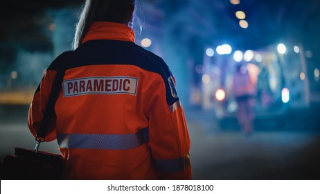Female EMS Paramedic Proudly Standing With Her Back Turned to Camera in High Visibility Medical Orange Uniform with "Paramedic" Text Logo. Successful Emergency Medical Technician or Doctor.