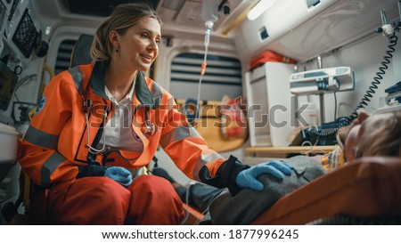 Female EMS Professional Paramedic Comforting Injured Patient on the Way to Hospital. Emergency Medical Care Assistant Puts Her Hand on Vinctim's Shoulder in a Friendly Way in an Ambulance.