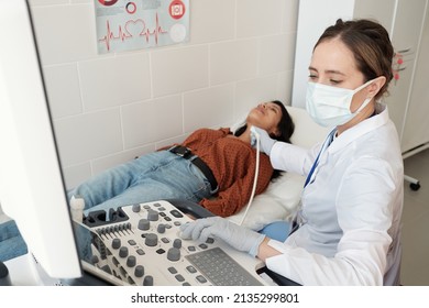 Female Endocrinologist In Whitecoat And Mask Sitting By Ultrasound Equipment During Examination Of Hispanic Patient On Couch