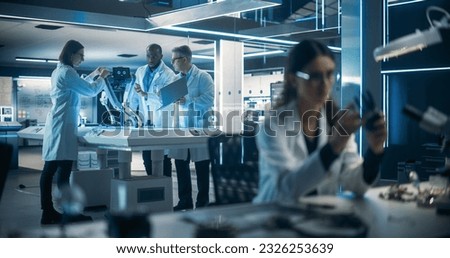 Female Engineer Fixing a High-Tech Bionic Hand Component. On Background Proffesional Industrial Robotics Specialists in Lab Coats Working on a Mobile AI Robot in a Tech Factory Facility.