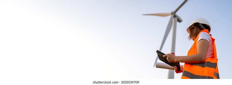 Female engineer in the helmet controls and maintains the operation of a wind turbine using a tablet on the background of windmill. Banner of production of alternative and renewable energy concept with - Shutterstock ID 2033539709