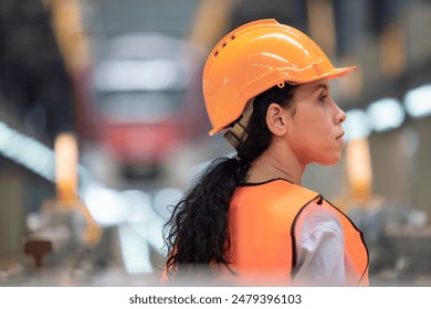 Female engineer with orange hard hat and safety vest is seen from behind, observing train inside maintenance depot. Highlights industrial work, safety, role of women in engineering and transportation - Powered by Shutterstock