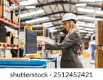 Female engineer, project manager standing in modern industrial factory by computer, making phone call. Team management in manufacturing facility. Side view.