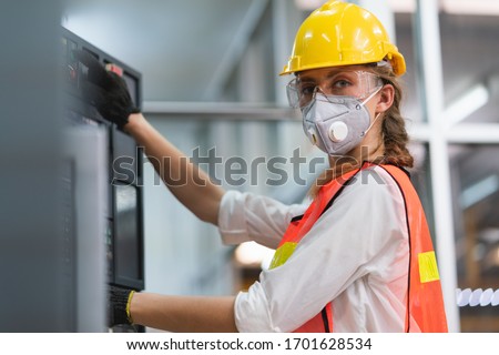 Female Engineer wear face mask with safety vest and yellow helmet operating control CNC Machinery at factory Industrial