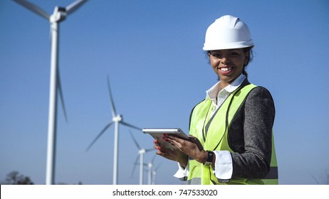 Female engineer wearing hard hat standing with digital tablet against wind turbine on sunny day and looking at camera smilingly