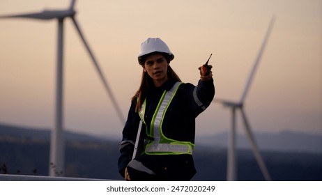 A female engineer wearing safety gear uses a radio to communicate while standing at a wind farm during sunset. - Powered by Shutterstock