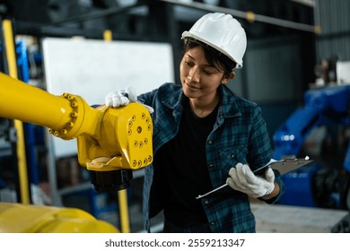 Female engineer wearing a safety helmet and gloves carefully examines a yellow robotic arm while taking notes on a clipboard in a high-tech industrial factory. - Powered by Shutterstock