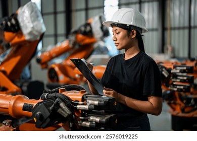 Female engineer wearing a white safety helmet and black shirt, standing in an industrial factory, analyzing orange robotic arms while taking notes on a clipboard.