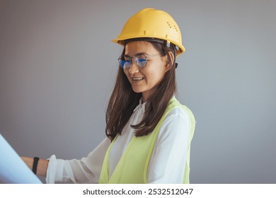 A female engineer wearing a yellow hard hat and safety vest reviews blueprints with focus and confidence in a professional office environment. - Powered by Shutterstock