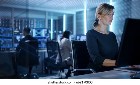 Female IT Engineer Works On Her Desktop Computer In Government Surveillance Agency. In The Background People At Their Workstations With Multiple Screens Showing Graphics.