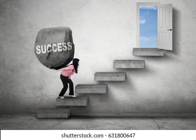 Female entrepreneur carries a boulder with Success word on her back while walking on the stairs toward a door
