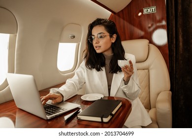 Female entrepreneur flies to a business meeting on a private plane, drinks coffee from a white cup and prints a document with a commercial offer on her laptop