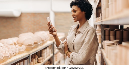 Female entrepreneur reading a text message on her smartphone. Happy businesswoman managing a store with handmade ceramic products. Creative female ceramist running a successful small business. - Powered by Shutterstock