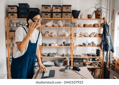 A female entrepreneur is standing at ceramics workshop and talking on the phone with customer while taking an order. A female potter is working with clay and having a phone conversation. Craftsmanship - Powered by Shutterstock