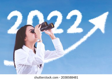 Female entrepreneur using binoculars while looking at clouds shaped 2022 numbers and upward arrow in the sky