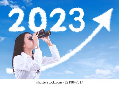 Female entrepreneur using binoculars while looking at clouds shaped 2023 numbers and upward arrow in the sky