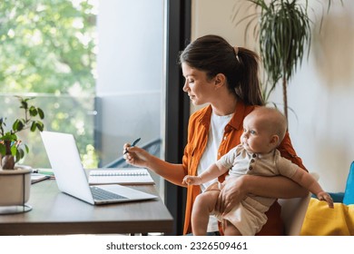 Female entrepreneur writing in notes while working on laptop and taking care of her baby at home. - Powered by Shutterstock