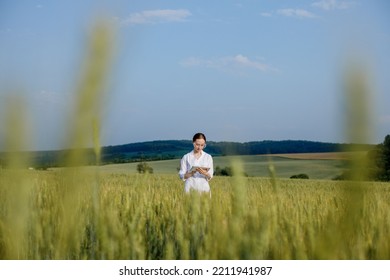 Female Environmentalist Scientist In A White Coat In A Green Field Of Unripe Ears Of Corn And Recording Data On A Tablet Device. Female Researcher, Biologist, Walking In The Field.