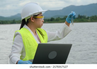 Female Environmentalist Wearing Gloves Takes Samples Of River Water To Examine Contaminants In Natural Waters And Record Data On A Laptop. Water And Ecology Concept.