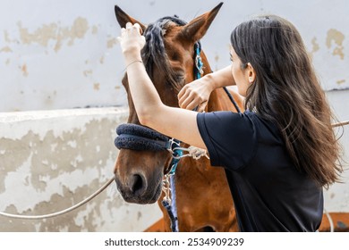 A female equestrian is seen carefully braiding the mane of a brown horse at a stable. The horse is tied securely, and the rider is focused on grooming, showcasing attention to detail and care. - Powered by Shutterstock