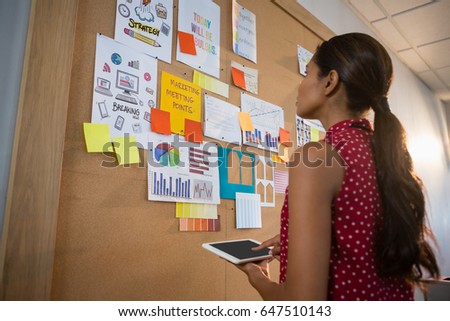 Female executive using digital while looking at the bulletin board in office