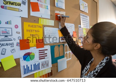 Female executive writing on sticky note at bulletin board in office