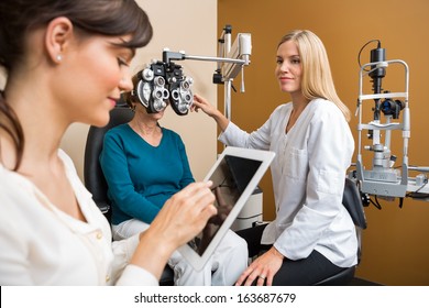 Female Eye Doctor Examining Senior Woman's Eyes While Colleague Using Digital Tablet In Store