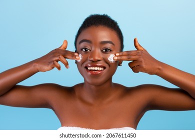 Female Facial Skincare. African American Lady Applying Moisturizer Cream On Cheeks, Posing Over Blue Studio Background, Smiling At Camera. Young Woman Moisturizing Skin