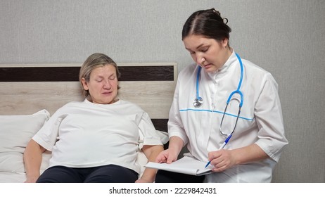 Female family doctor makes notes on clipboard listening to complaints of lady patient. Mature woman lies in bed talking to experienced therapist at home - Shutterstock ID 2229842431
