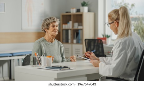 Female Family Doctor Is Sitting Behind A Desk And Speaking To A Senior Woman During Consultation In A Health Clinic. Physician In Lab Coat Prescribing Drugs To Elderly Patient In Hospital Office.