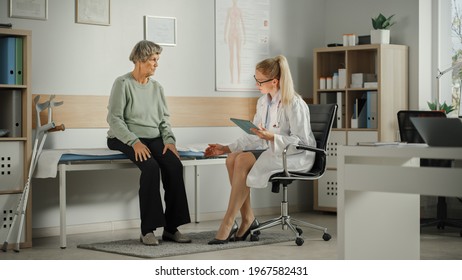 Female Family Doctor Speaking To A Senior Woman During Consultation In Health Clinic And Asking About Her Leg Injury. Physician In Lab Coat Suggesting Treatment To Elderly Patient In Hospital Office.