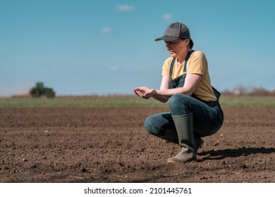 Female farmer agronomist checking the quality of ploughed field soil before sowing season, agricultural tractor in the background, selective focus