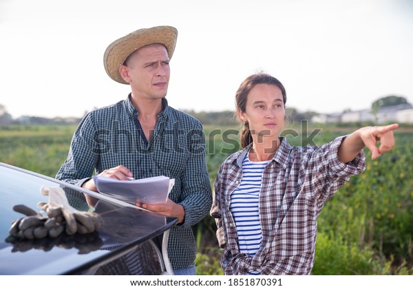 Female farmer discussing\
papers with her partner while standing near car on farm field on\
autumn day