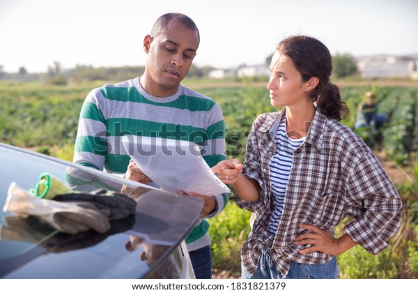 Female farmer discussing some\
papers with hispanic partner while standing near car on farm\
field