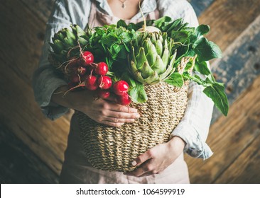 Female farmer in linen apron holding basket of fresh garden vegetables and greens in her hands, rustic wooden barn wall at background, horizontal composition. Local market or organic produce concept