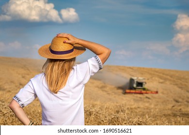 Female Farmer Looking At Combine Harvester At Field. Woman With Straw Hat Examining Harvest Of Organic Farm. Agriculture Concept