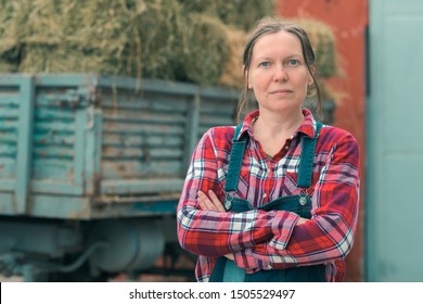 Female farmer posing in front of hay wagon. Portrait of woman farm worker in plaid shirt and bib overalls by the tractor trailer filled with dairy farm livestock feed hay bales.