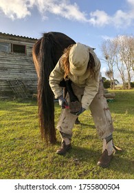 A female farrier working on a horse's hoof with a hoof pick. A barn and green grass in the background.